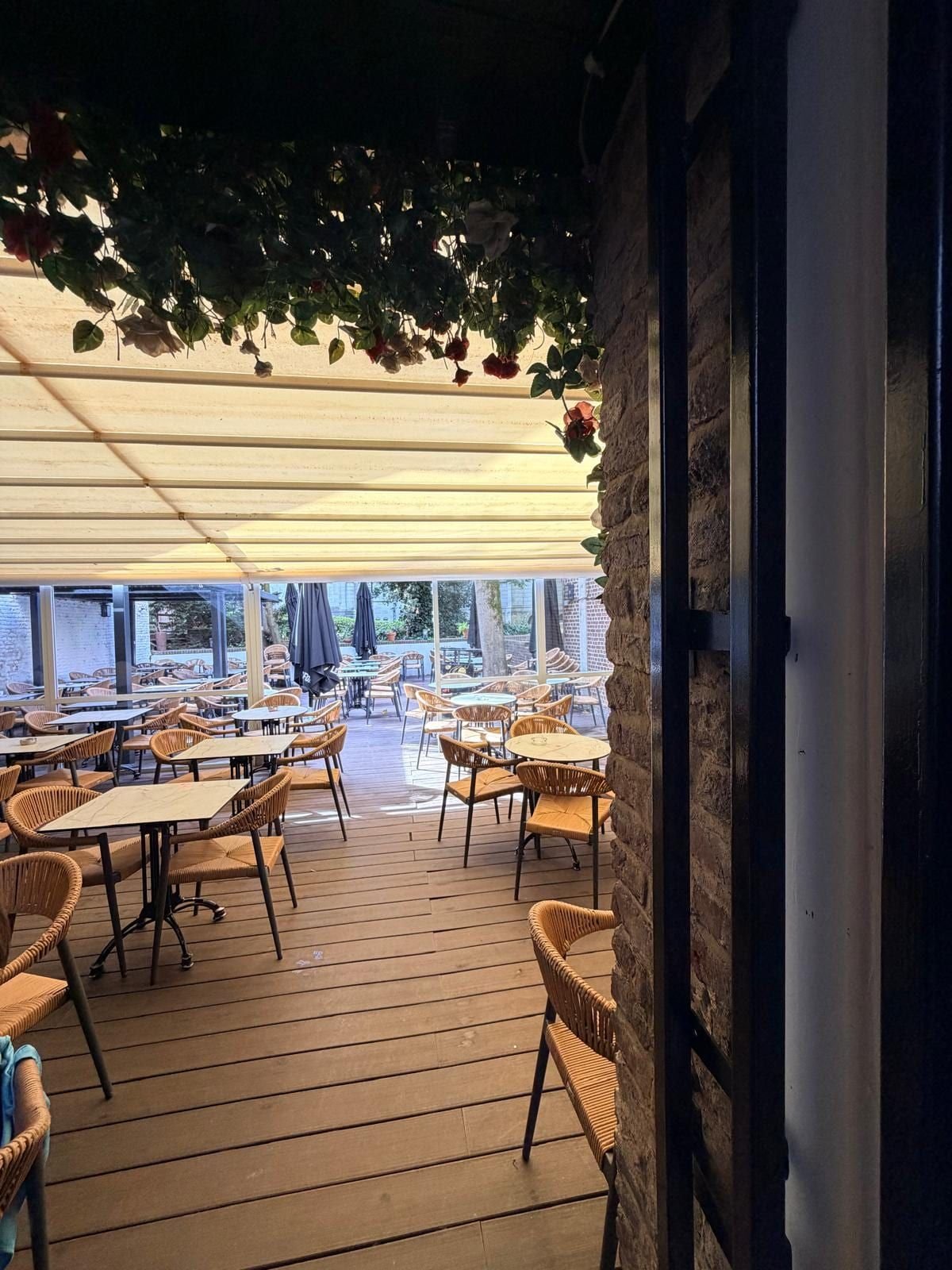 La partie couverte de la terrasse vue depuis la salle, sous une véranda et des fleurs suspendues